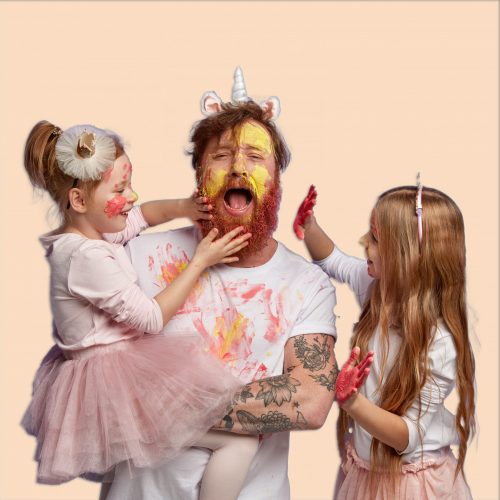 Photo of upset tired dad being smeared with watercolors by children who leave palm prints on his face, has tattooed arms, poses with daughters against purple background. Girs play with colors, paints party_komunia_bez_animatora_jakie_zabawy_rozrywka_bez_animatorów